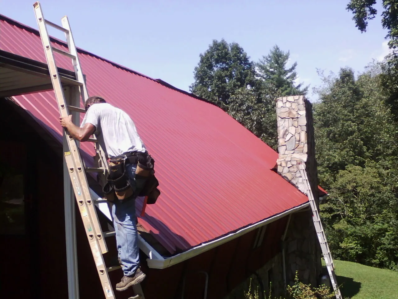 Roofer on a ladder beside a red metal roof and stone chimney — Soffit & Fascia Repair by Roofing Brothers in Keystone